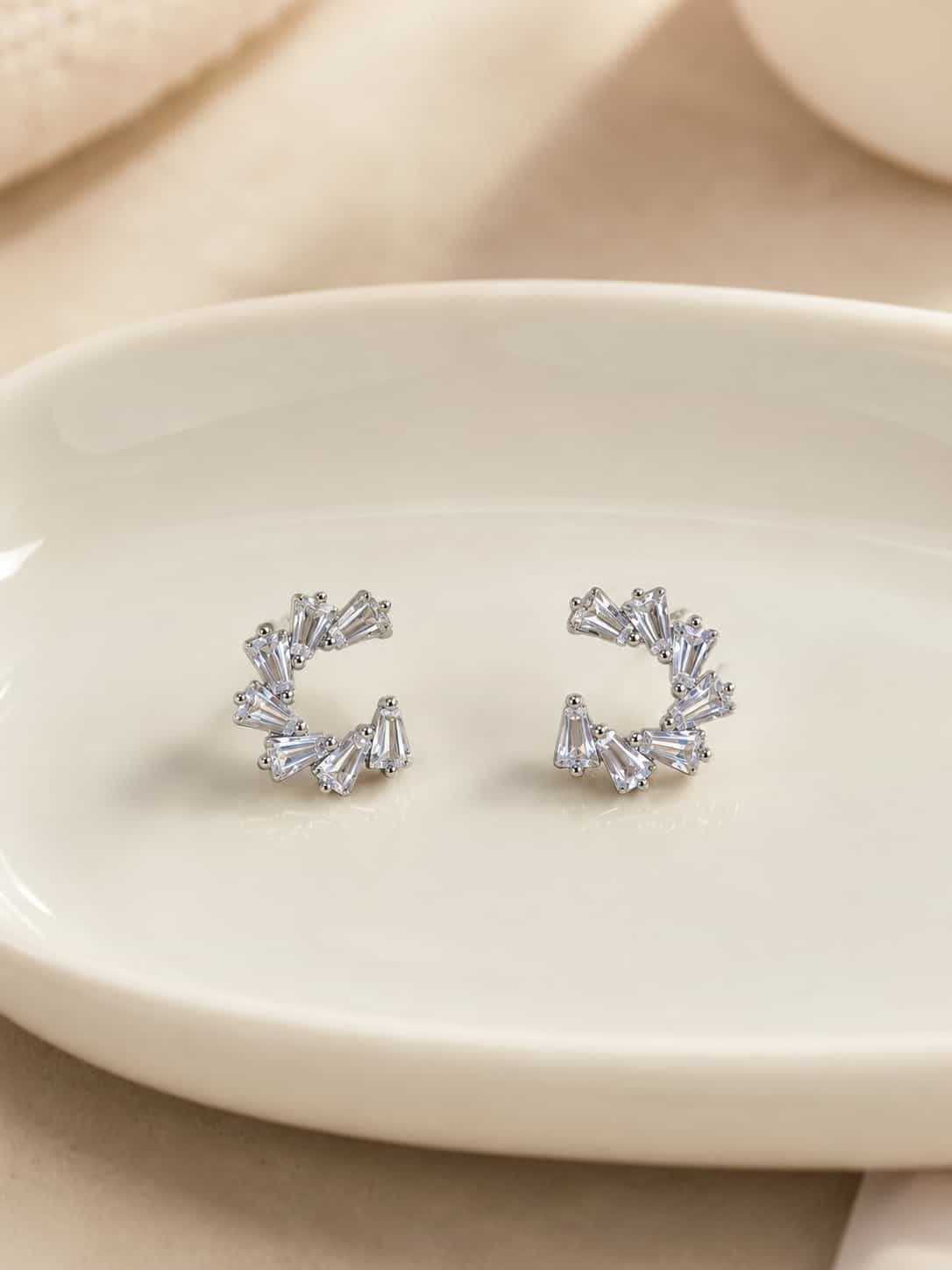 Platinum polished crystal fan open hoop earrings with tapered clear stones on a cream ceramic display