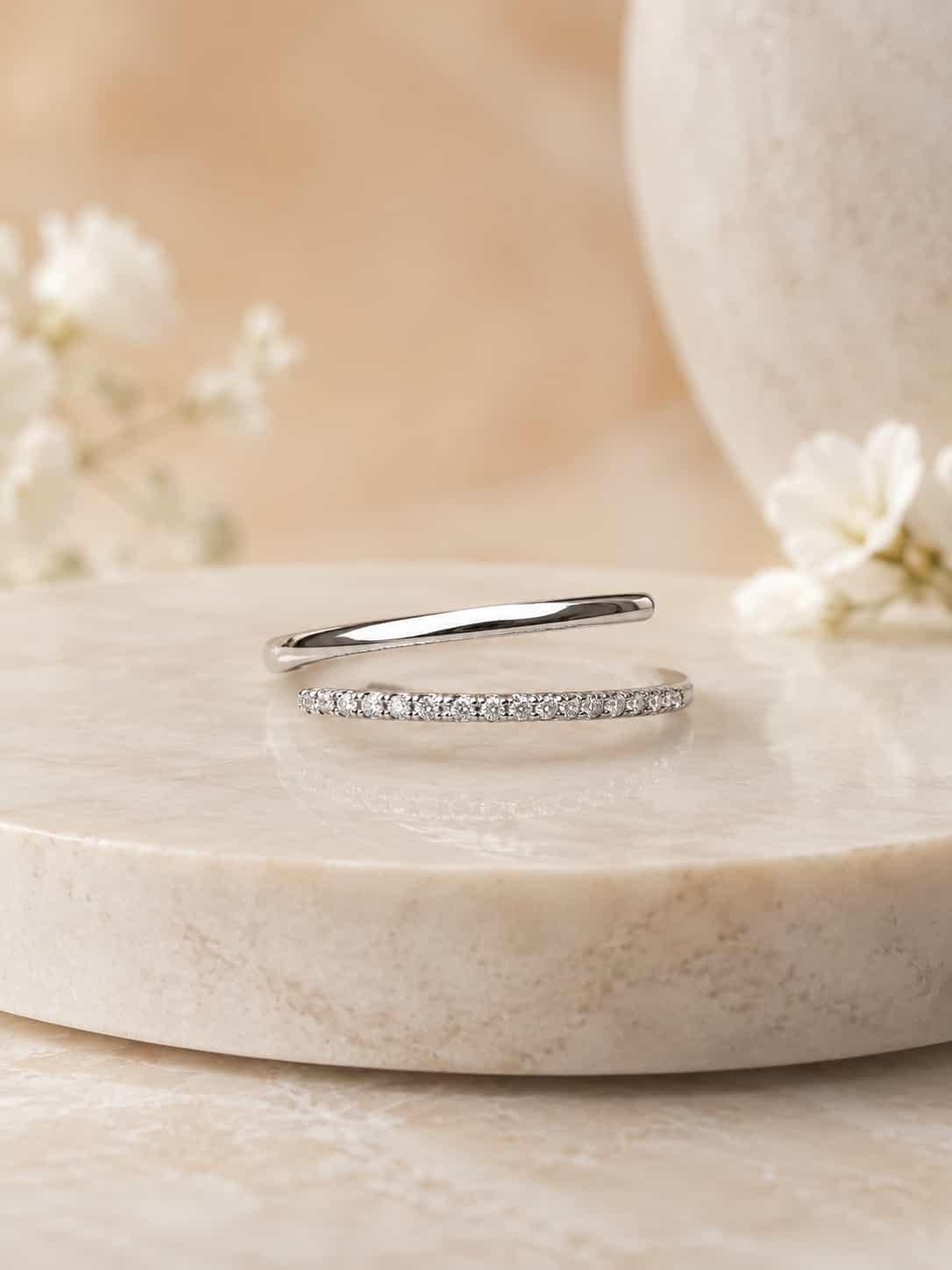 Naira double-row open wrap ring with polished and clear-stone bands on a cream stone plinth beside white flowers