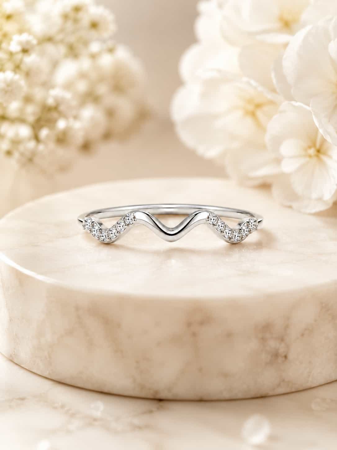 Serena platinum-polished wave ring displayed on a white marble pedestal with soft ivory flowers