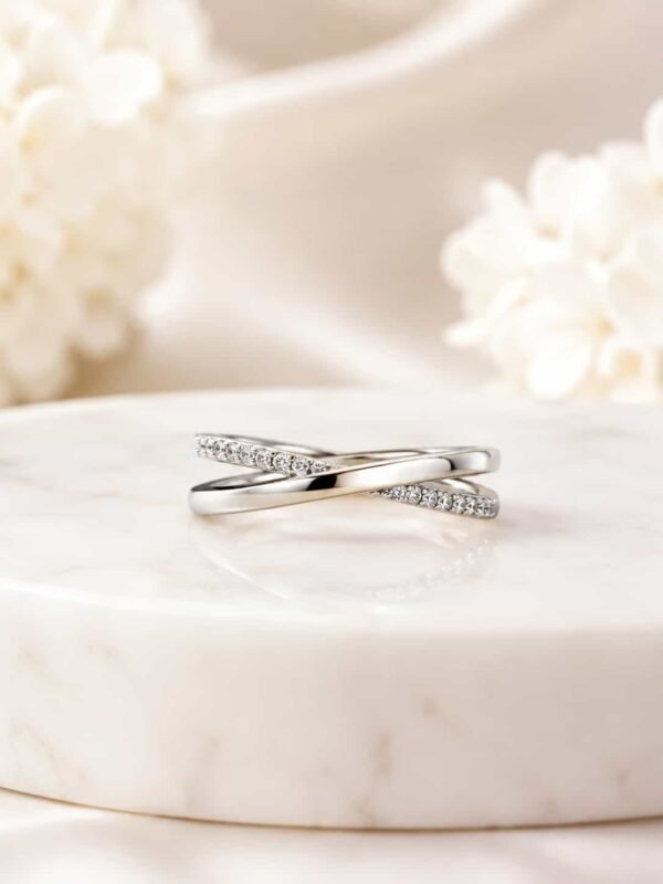 Elara platinum-polished criss-cross ring displayed on a white marble pedestal with soft ivory flowers