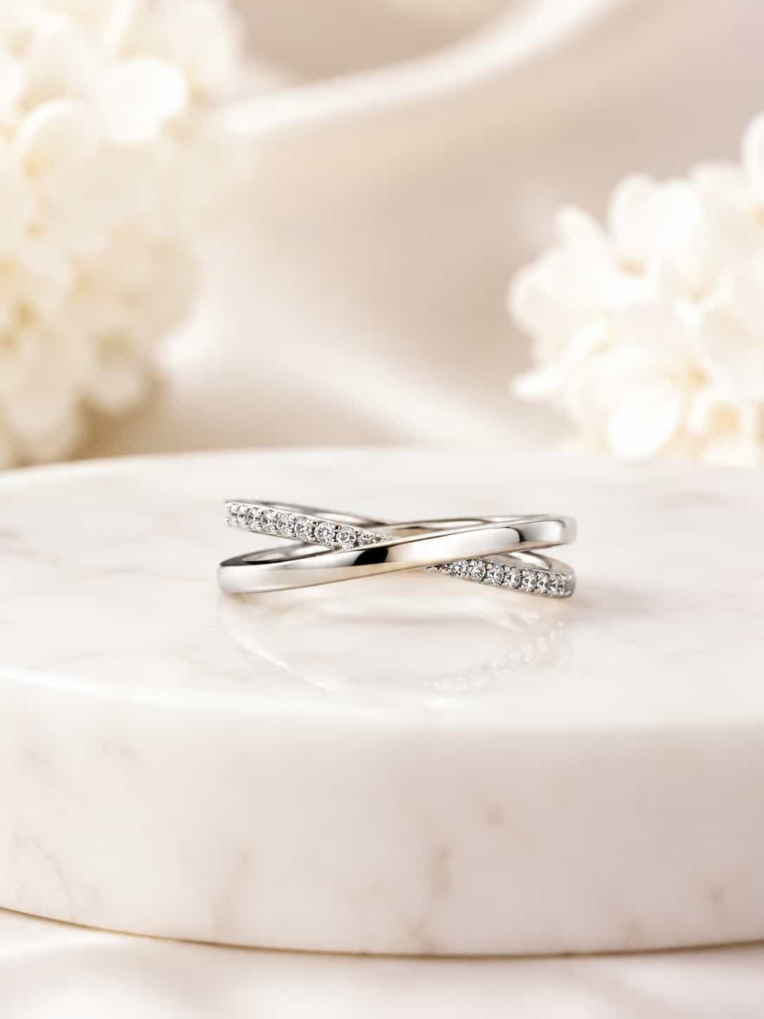 Elara platinum-polished criss-cross ring displayed on a white marble pedestal with soft ivory flowers
