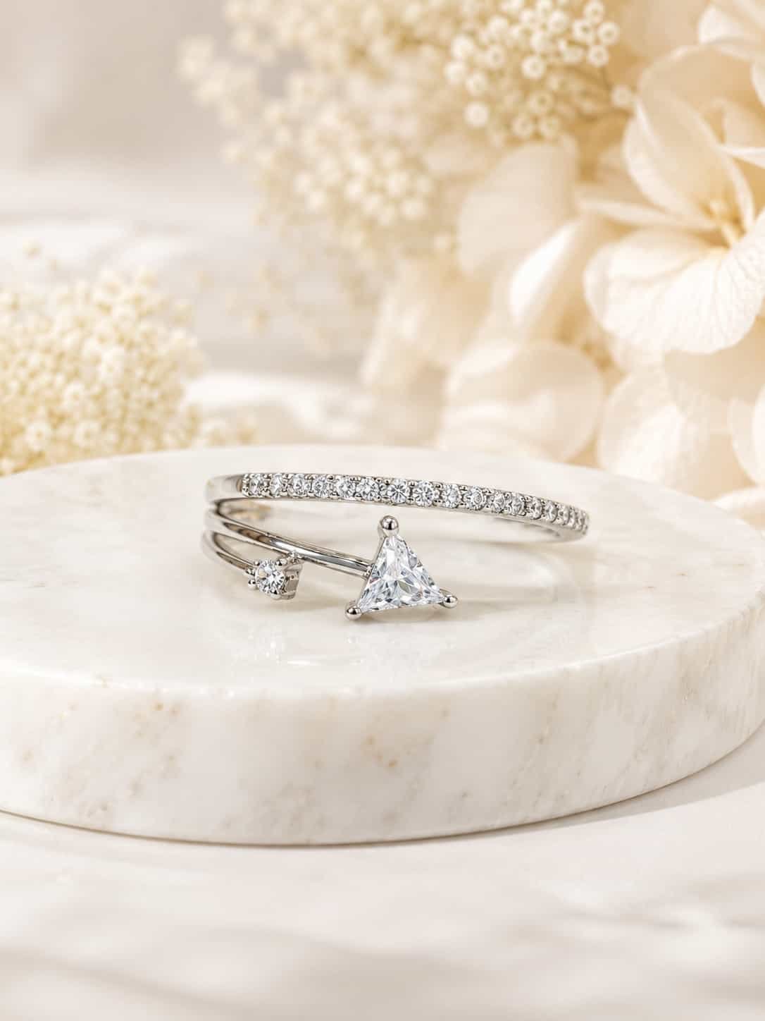 Evara platinum-polished triangle open ring displayed on a white marble pedestal with soft ivory flowers