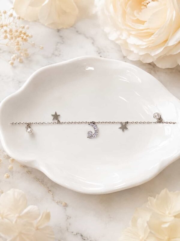 Delicate celestial bracelet with crescent moon, star and pearl-style charms displayed on white jewellery dish